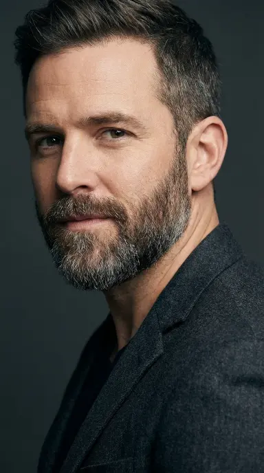 A close-up editorial photograph of a stylish man in his late 30s with a neatly groomed medium-length beard